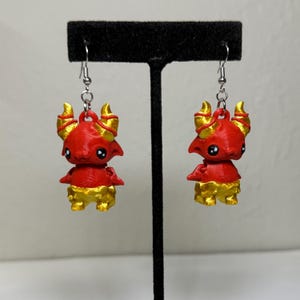 May include: A pair of red and gold devil-themed earrings. Each earring features a small, cartoonish devil figure with horns, wings, and a tail. The devil's body is red, with gold accents on the horns, wings, and lower body. The earrings hang from silver-colored hooks.