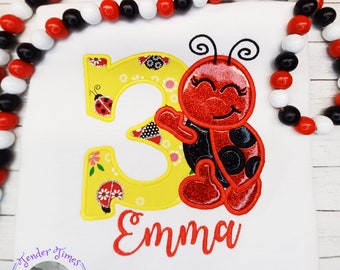 May include: White t-shirt featuring a smiling ladybug and the number 3 in yellow with floral accents. The name "Emma" is written in red script below the number. A beaded garland frames the shirt.