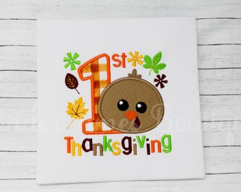 May include: White t-shirt with an embroidered Thanksgiving design. The design features a cartoon turkey head, the number 1 in orange plaid, and the words "1st Thanksgiving" in colorful letters, with fall leaves.