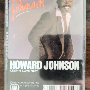 Può includere: Una musicassetta vintage dell'album "Keepin' Love New" di Howard Johnson. La copertina presenta una foto di Howard Johnson in giacca e cravatta con una camicia con colletto rosa. Il titolo dell'album e il nome dell'artista sono in arancione e bianco.
