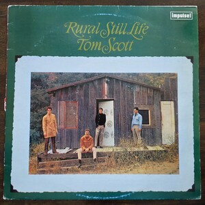 Może przedstawiać: Vintage okładka albumu winylowego "Rural Still Life" Toma Scotta. Okładka przedstawia zdjęcie drewnianej chaty z czterema osobami na zewnątrz. Tytuł albumu i nazwisko artysty są w ozdobnej czcionce.