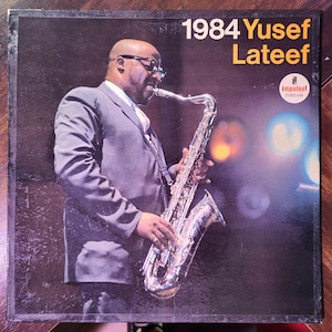 Puede incluir: Portada del álbum "1984" de Yusef Lateef. La portada presenta una fotografía de un hombre con traje tocando el saxofón. El título del álbum y el nombre del artista están en texto blanco y amarillo. El logotipo de Impulse! Records es visible.