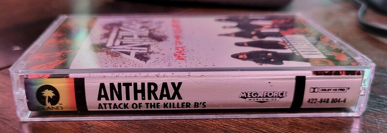 May include: A clear cassette tape featuring the band Anthrax's album "Attack of the Killer B's." The album cover shows the band members and the text "Anthrax" in bold letters. The cassette case has a rainbow-colored Island Records logo.