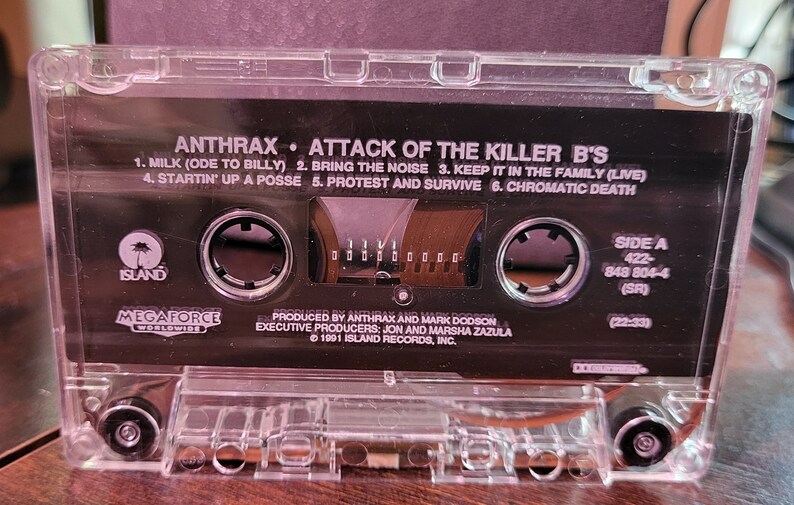 May include: Clear cassette tape with the album title "Attack of the Killer B's" by Anthrax. The black label features song titles and the Island Records logo. The tape is in a clear plastic case.