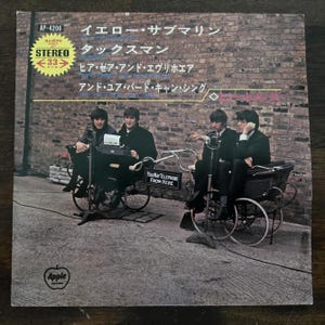 May include: Album cover for "Yellow Submarine" by The Beatles. The image features the band members seated in antique-style vehicles against a brick wall. Japanese text and the Apple Records logo are visible.