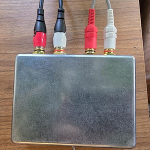 Puede incluir: Una peque&ntilde;a caja met&aacute;lica rectangular de color plateado con cuatro entradas de cable de audio. Los cables son negros, rojos y blancos, con conectores dorados. Un peque&ntilde;o interruptor est&aacute; situado en la parte inferior de la caja.