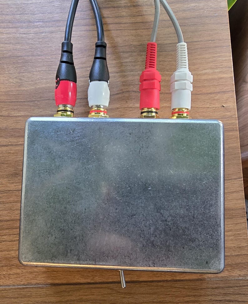 Puede incluir: Una peque&ntilde;a caja met&aacute;lica rectangular de color plateado con cuatro entradas de cable de audio. Los cables son negros, rojos y blancos, con conectores dorados. Un peque&ntilde;o interruptor est&aacute; situado en la parte inferior de la caja.