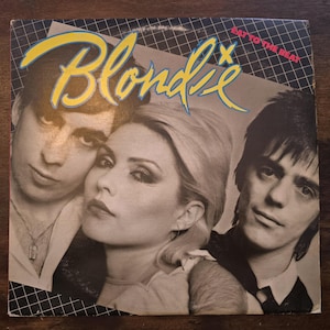 May include: Album cover for Blondie's "Eat to the Beat." The cover features three band members with the band's name in yellow, stylized text. The album title is in pink text. The background has a grid pattern.