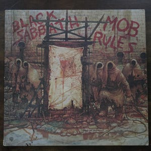 May include: Album cover art for Black Sabbath's "Mob Rules." The artwork features a brick wall with the band's name and album title in red graffiti. Figures in hooded cloaks stand near a tattered cloth, with a dark, ominous atmosphere.