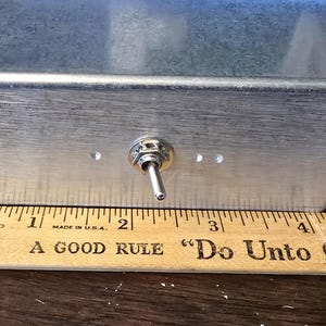 Puede incluir: Una caja rectangular de metal cepillado con un interruptor de palanca y tres peque&ntilde;os agujeros. Una regla de madera con el texto "A GOOD RULE 'Do Unto Others As" est&aacute; colocada debajo. La caja mide aproximadamente 10 cm de ancho.