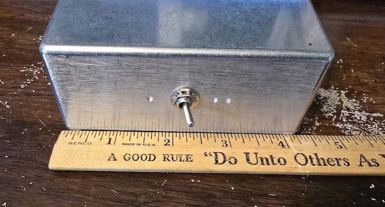 Puede incluir: Una caja rectangular de metal cepillado con un interruptor de palanca y tres peque&ntilde;os agujeros. Una regla de madera con el texto "A GOOD RULE 'Do Unto Others As" est&aacute; colocada debajo. La caja mide aproximadamente 10 cm de ancho.