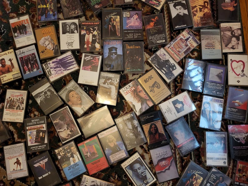 May include: A selection of vintage cassette tapes, displaying album artwork with text and images. The tapes are arranged on a patterned surface, representing a range of music genres and artists from different periods.