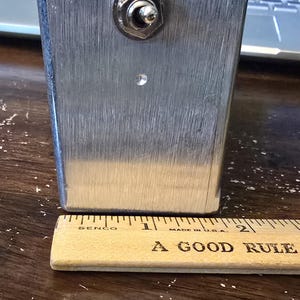Puede incluir: Un objeto rectangular de metal cepillado con un interruptor basculante en la parte superior y un peque&ntilde;o agujero debajo. Una regla de madera con el texto "A GOOD RULE" y marcas en pulgadas est&aacute; en la parte inferior. El objeto mide unos 5 cm de altura.