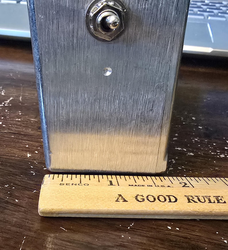 Puede incluir: Un objeto rectangular de metal cepillado con un interruptor basculante en la parte superior y un peque&ntilde;o agujero debajo. Una regla de madera con el texto "A GOOD RULE" y marcas en pulgadas est&aacute; en la parte inferior. El objeto mide unos 5 cm de altura.