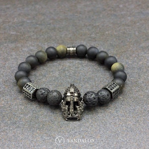 May include: A black and grey beaded bracelet with a silver helmet charm. The bracelet features black lava rock beads and grey agate beads.
