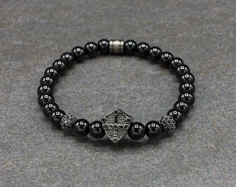 May include: Black onyx beaded bracelet with a silver shield charm featuring a cross design. The bracelet has a silver clasp.