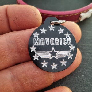 May include: Black metal dog tag with a silver star and wings design. The tag says "Maverick" in silver lettering and is surrounded by silver stars.
