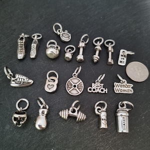 May include: A collection of silver charm pendants with fitness and workout themes. The charms include a dumbbell, a weight plate, a kettlebell, a running shoe, a boxing glove, a shaker bottle, and a charm that says "Best Coach".