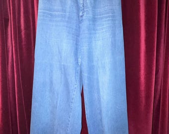 May include: High-waisted blue denim trousers featuring a wide-leg silhouette, a central button closure, and two small horizontal pockets located just below the waistband. The garment is suspended from a metal hanger.