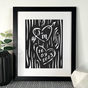 May include: A framed art print with a black frame and white mount. The black artwork features two white hearts, one with initials and an arrow, the other with a date. The frame is black.