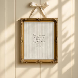 May include: A framed print with a gold ornate frame and a cream-colored background. The print features the text "Fixing our eyes on Jesus, the author and perfecter of our faith. Hebrews 12:2." A cream-colored ribbon is tied at the top.
