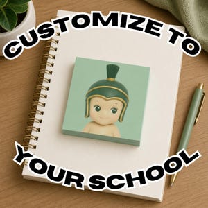 Puede incluir: Un bloc de notas verde claro con una figura de dibujos animados con un casco verde, sobre un cuaderno de espiral. También se ve un bolígrafo verde y las palabras "CUSTOMIZE TO YOUR SCHOOL".