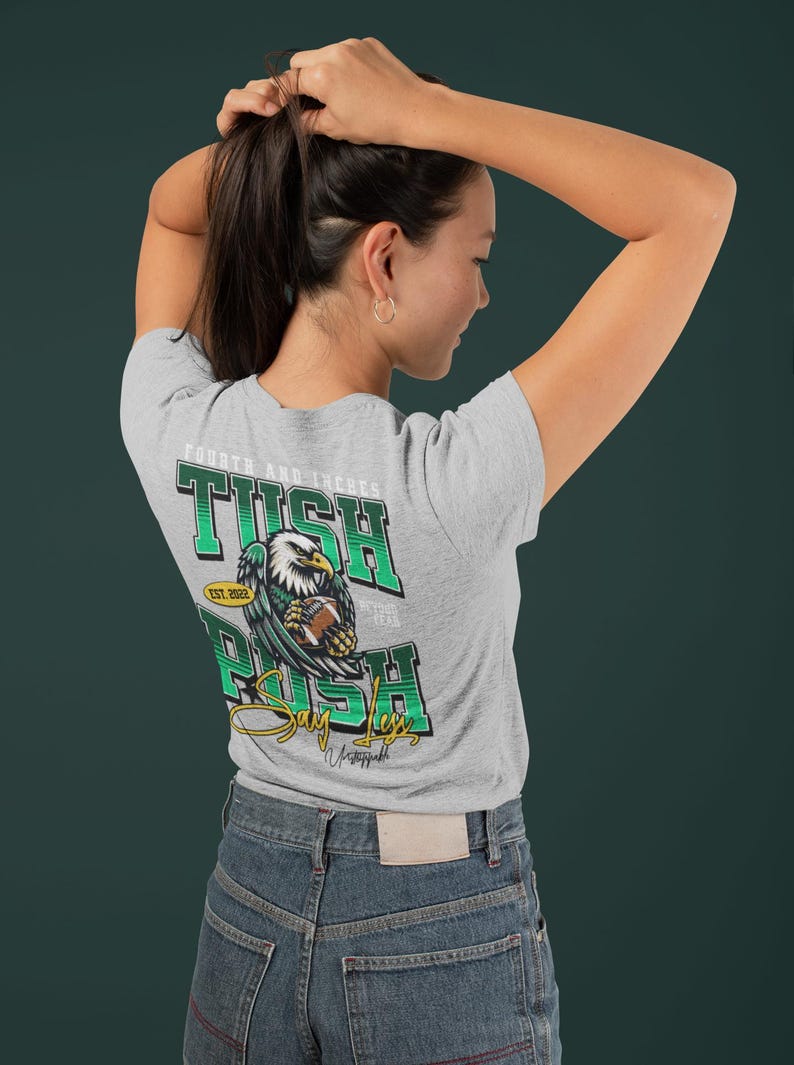May include: Grey t-shirt with a green and white graphic on the back. The graphic features an eagle holding a football, with the words "TUSH RUSH" and "Say Less" in a stylized font. The text "EST. 2022" is also included.