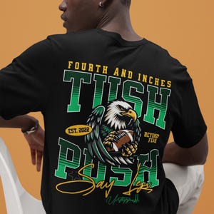 May include: Black t-shirt with a football-themed graphic. The design features an eagle clutching a football, with text that reads "Fourth and Inches", "TUSH PUSH", "Say Legs", and "Beyond Fear". The shirt includes the year "Est. 2022".