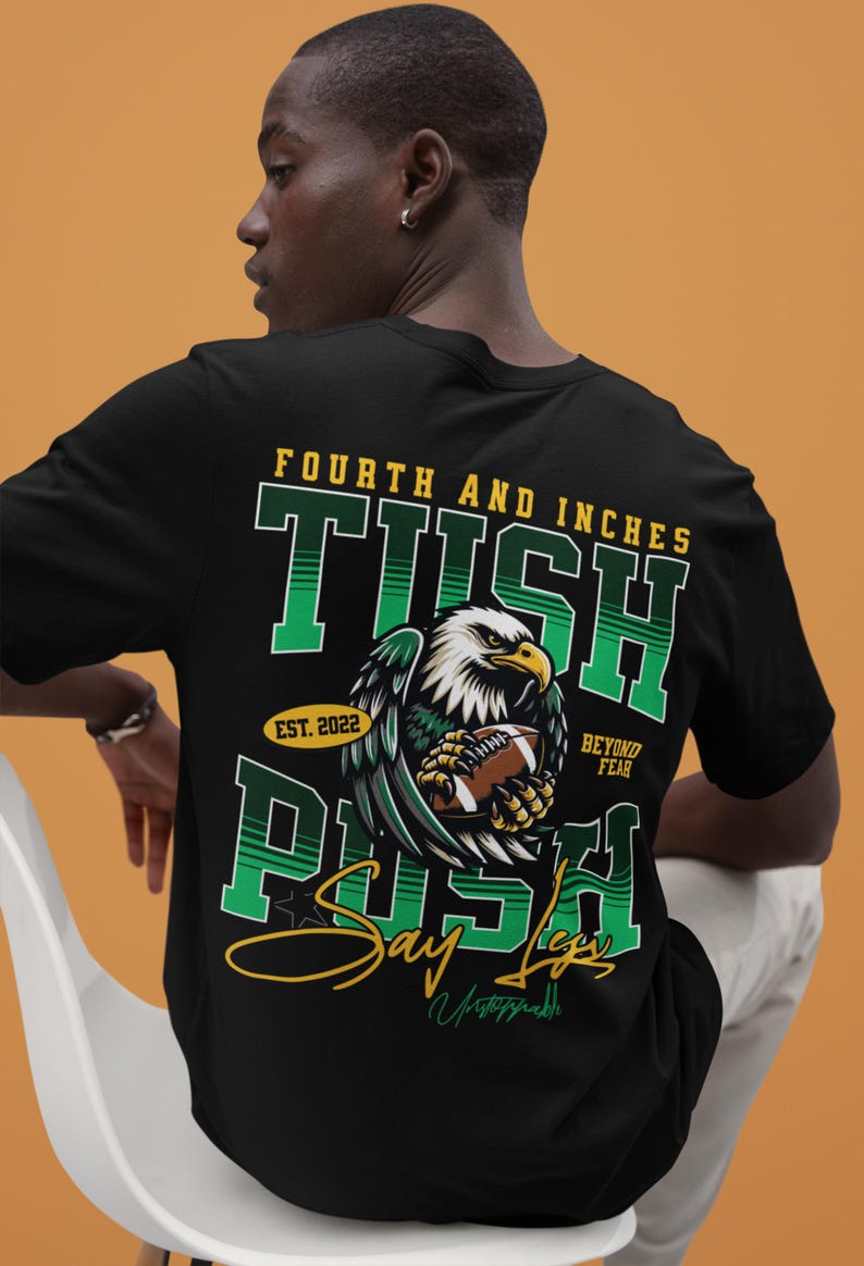 May include: Black t-shirt with a football-themed graphic. The design features an eagle clutching a football, with text that reads "Fourth and Inches", "TUSH PUSH", "Say Legs", and "Beyond Fear". The shirt includes the year "Est. 2022".