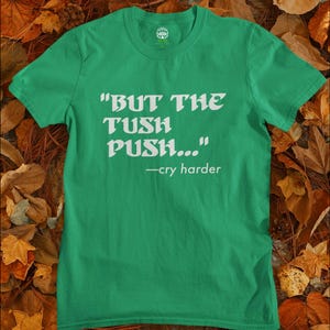 May include: A green t-shirt with the text "BUT THE TUSH PUSH..." and "-cry harder" in white. The shirt is displayed on a bed of autumn leaves.