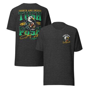 May include: Dark grey t-shirts with a football theme. One shirt features a large graphic of an eagle holding a football, with the text "FOURTH AND INCHES TUSH PUSH Say Less" and "EST. 2022". The other shirt has a smaller eagle graphic and the text "TUSH PUSH Unstoppable".