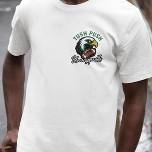 May include: White t-shirt featuring a green and black eagle graphic holding a football. The text "Tush Push Unstoppable" is above and below the graphic. The shirt has a crew neck.