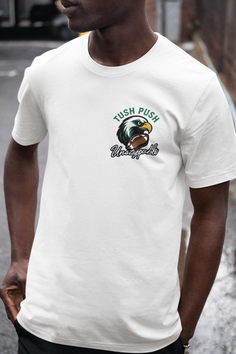 May include: White t-shirt featuring a green and black eagle graphic holding a football. The text "Tush Push Unstoppable" is above and below the graphic. The shirt has a crew neck.