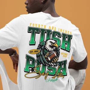 May include: White t-shirt with a green and yellow graphic. The design shows an eagle clutching a football, with text reading "Fourth and Inches", "Tush Rush", and "Say Less". The shirt also includes "Est. 2022" and "Beyond Fear".