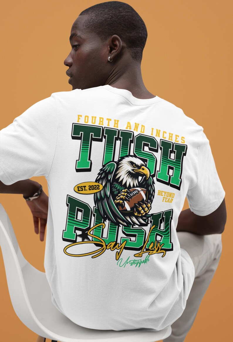 May include: White t-shirt with a green and yellow graphic. The design shows an eagle clutching a football, with text reading "Fourth and Inches", "Tush Rush", and "Say Less". The shirt also includes "Est. 2022" and "Beyond Fear".