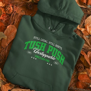 Puede incluir: Una sudadera con capucha verde oscuro con el texto "TUSH PUSH Unstoppable Since Day One" en blanco y verde. La sudadera tiene una capucha con cordón y un bolsillo delantero. El texto "Still Legal. Still Hurts." está encima del texto principal.