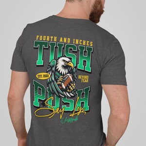 May include: Charcoal grey t-shirt with a football-themed design. The graphic includes an eagle clutching a football, with text reading "Fourth and Inches," "Tush Push," and "Say Less." The shirt also features "Est. 2022" and "Beyond Fear."