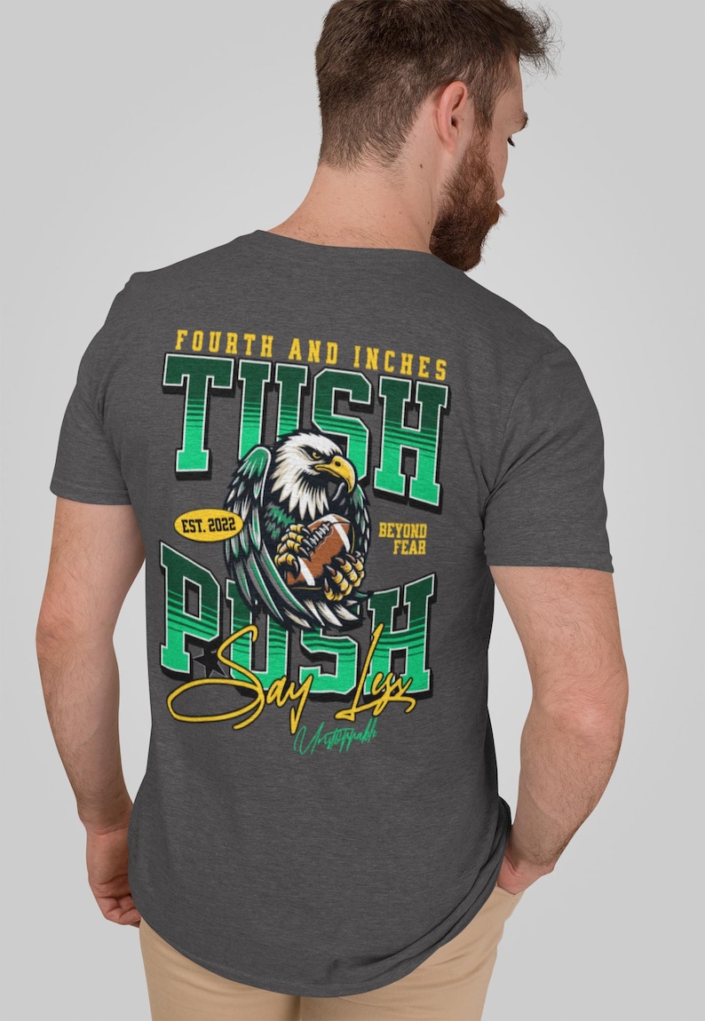 May include: Charcoal grey t-shirt with a football-themed design. The graphic includes an eagle clutching a football, with text reading "Fourth and Inches," "Tush Push," and "Say Less." The shirt also features "Est. 2022" and "Beyond Fear."
