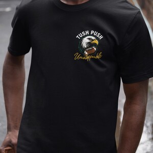 May include: Black t-shirt with an eagle head and football graphic, and the words "Tush Push Unstoppable" in white and gold. The shirt is made of a soft, comfortable material.