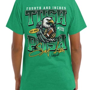 May include: Green t-shirt with a graphic design featuring an eagle clutching a football. The text on the shirt reads "FOURTH AND INCHES TUSH PUSH" with "EST. 2022" and "BEYOND FEAR" included. The shirt also has the words "Say Less" and "Unstoppable".