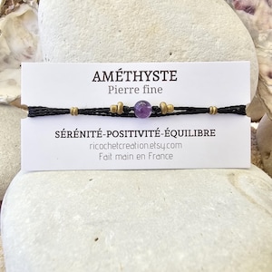 Peut inclure: Un bracelet en cordon noir avec des perles dorées et une pierre d'améthyste violette. Une carte blanche avec le texte "AMÉTHYSTE Pierre fine" et "SÉRÉNITÉ-POSITIVITÉ-ÉQUILIBRE" est devant le bracelet. Le fond est une pierre claire.