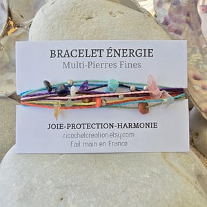 May include: A multi-coloured bracelet featuring various stones and beads. The bracelet is made with colourful threads and has a tag that reads "BRACELET ÉNERGIE Multi-Pierres Fines JOIE-PROTECTION-HARMONIE ricochetcreation.etsy.com Fait main en France".