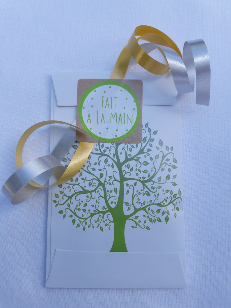 May include: A white envelope with a green tree design and a brown sticker that says "FAIT &Agrave; LA MAIN". The envelope is tied with a gold and silver ribbon.