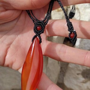 May include: A red carnelian gemstone pendant on a black cord necklace. The pendant is shaped like a leaf and has a smooth, polished finish. The necklace has two red beads at the ends of the cord.