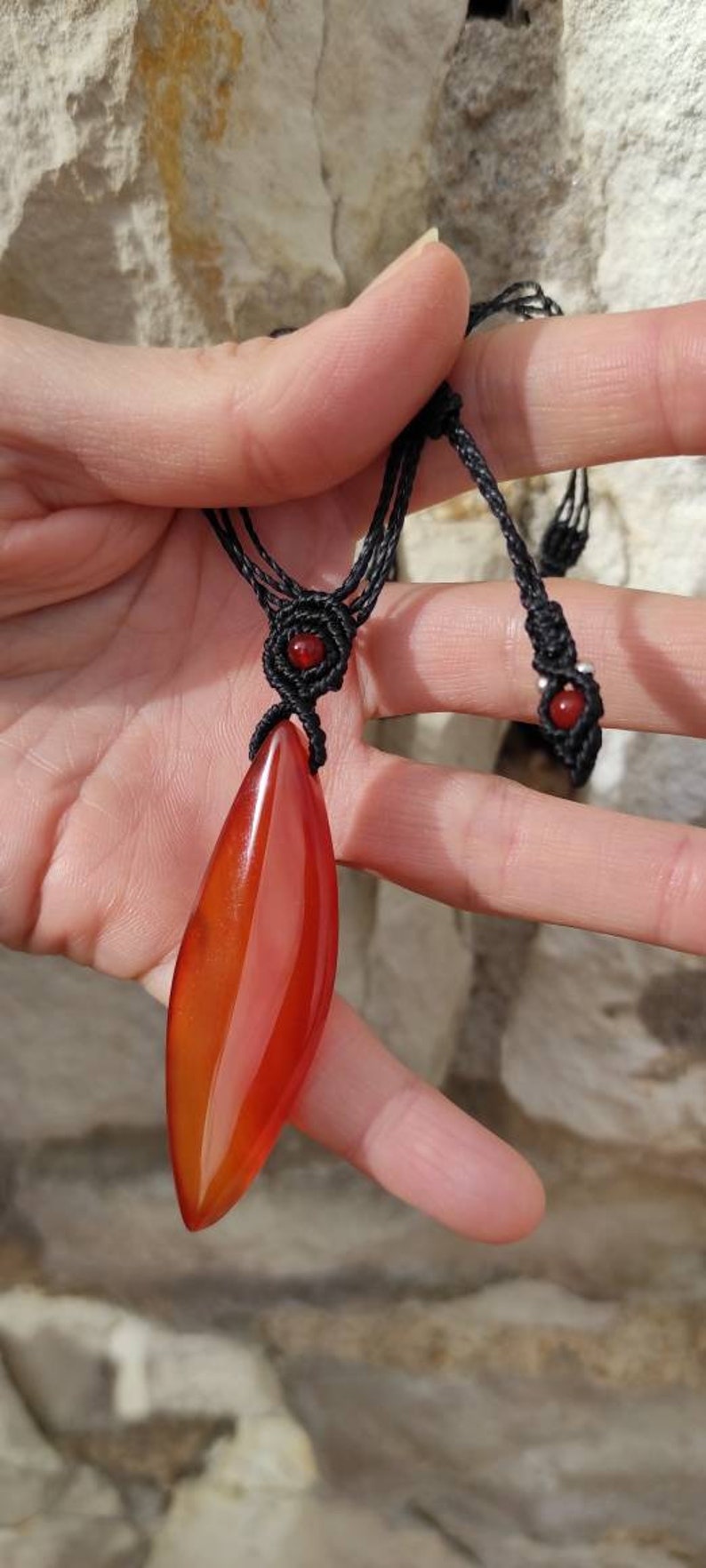 May include: A red carnelian gemstone pendant on a black cord necklace. The pendant is shaped like a leaf and has a smooth, polished finish. The necklace has two red beads at the ends of the cord.