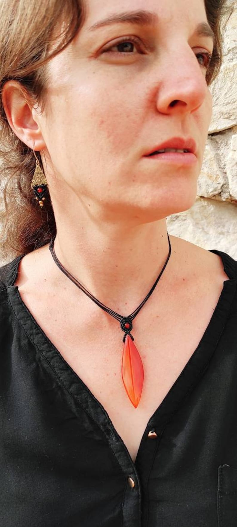 May include: A woman wearing a black shirt with a red and orange leaf pendant necklace. The necklace is made of black cord and has a small red bead at the top.