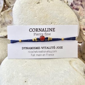 May include: A blue and gold bracelet with a red carnelian gemstone. The card reads "CORNALINE Pierre fine" and "DYNAMISME-VITALITÉ-JOIE ricochetcreation.etsy.com Fait main en France".