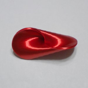 Puede incluir: Una sola flor de satén rojo, enroscada en forma de espiral. La flor está hecha de tela y tiene un acabado liso y brillante.