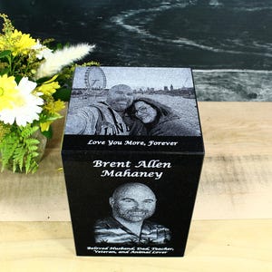May include: A black rectangular stone memorial urn featuring a laser-etched portrait of a man and a couple, with the text "Love You More, Forever," "Brent Allen Mahaney," and "Beloved Husband, Dad, Teacher, Veteran, and Animal Lover" engraved on it.