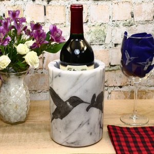 May include: White marble wine cooler with a hummingbird design. The cooler is holding a bottle of red wine.